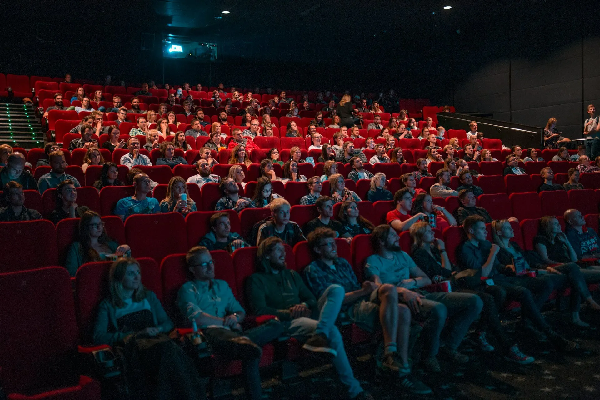 A full cinema audience in red seats watching a screening, faces lit by the light from the screen and the projector beam visible above the back rows