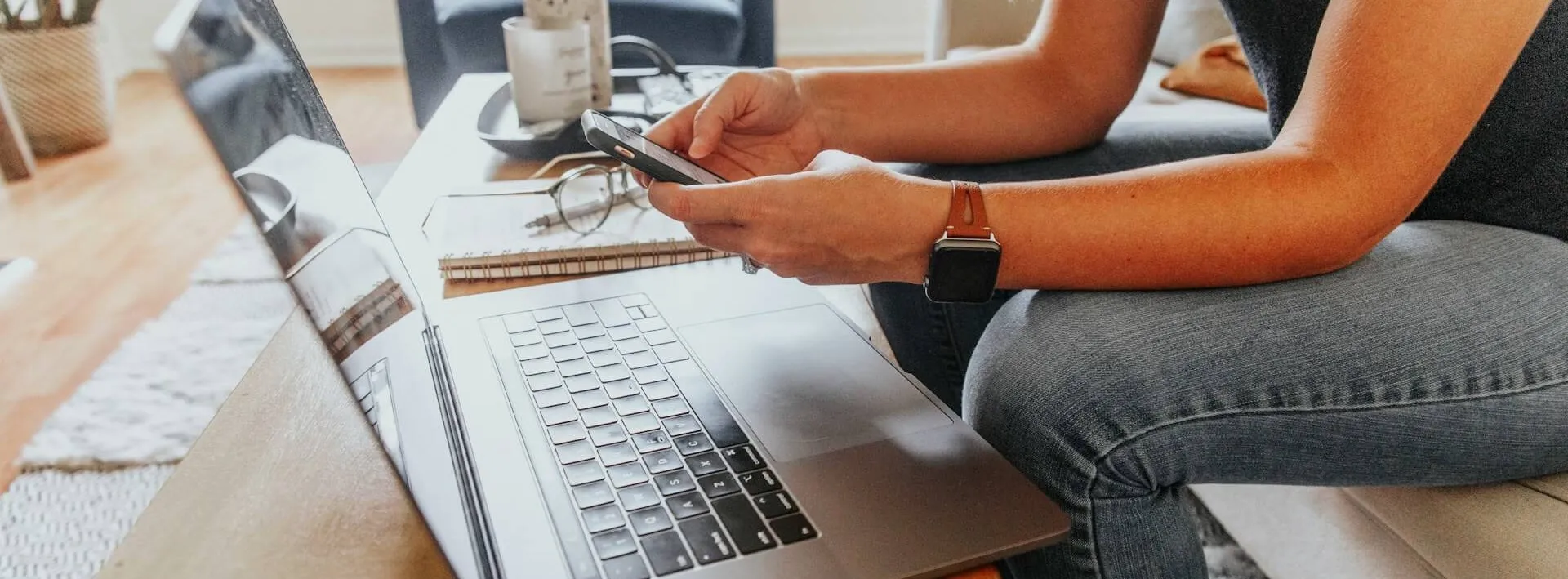 Someone sitting on a sofa holding a phone in both hands, with an open laptop on the coffee table in front of them, notebook and glasses beside it