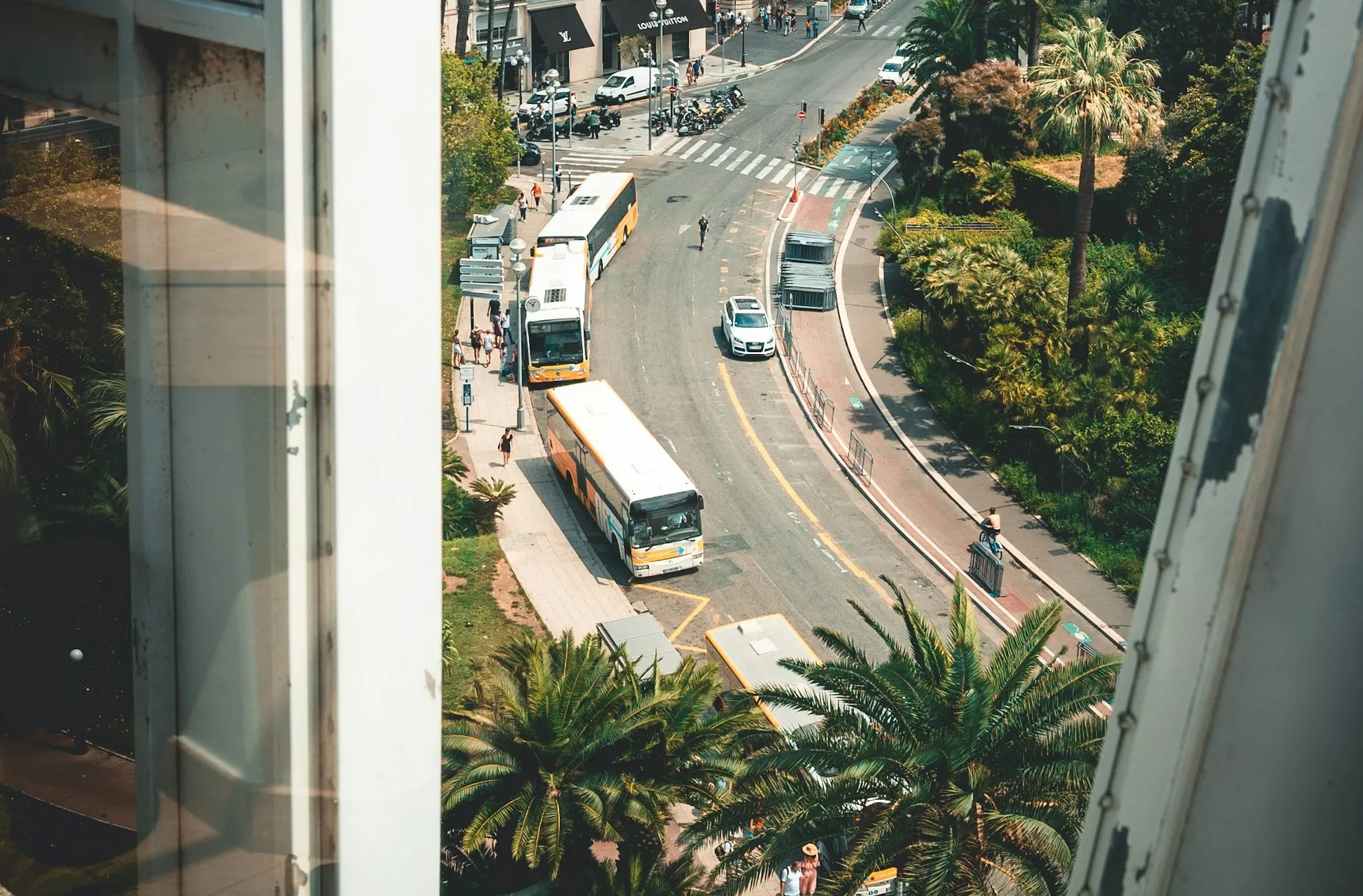 Two buses at a stop on a curving Nice street seen from a window above, with commuters waiting on the sidewalk, a cycle lane alongside and palms lining the road