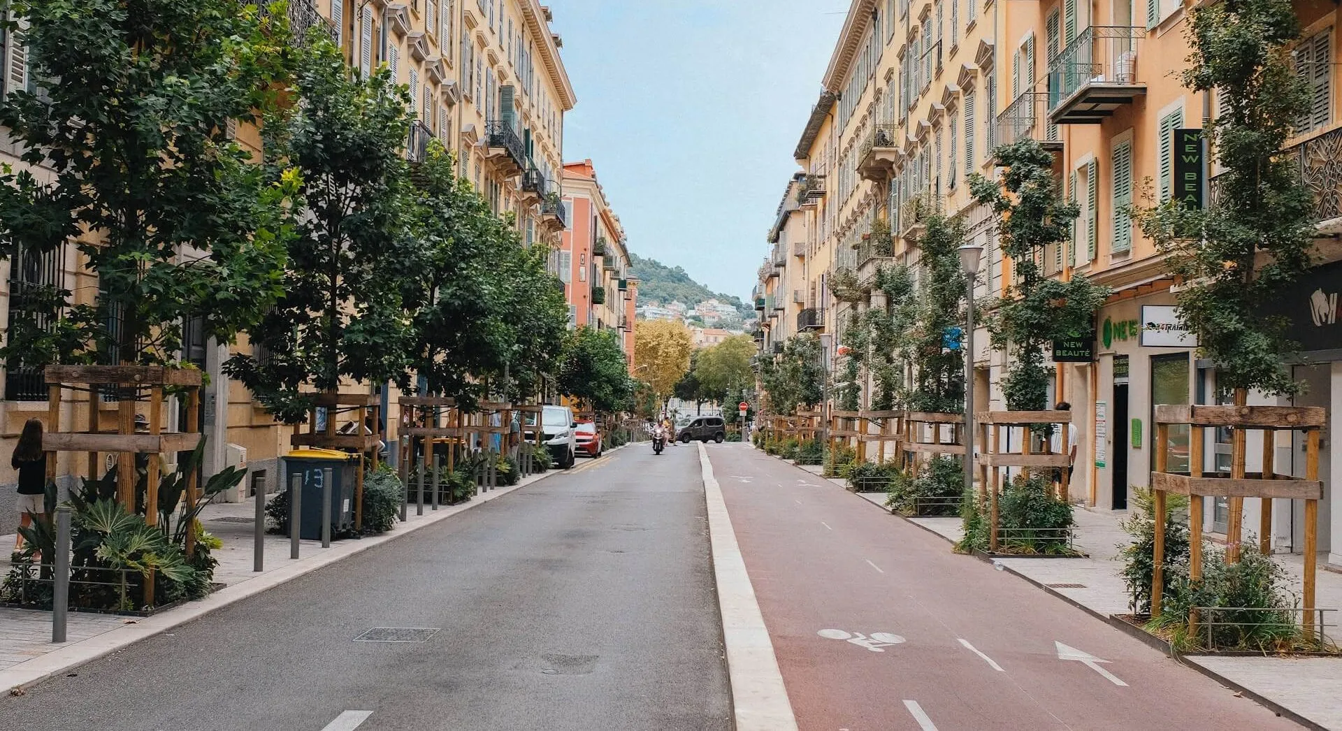A residential street in Nice lined with pastel apartment buildings, shuttered windows and wrought-iron balconies, young plane trees along both sidewalks and a cycle lane running toward the hills at the end of the street