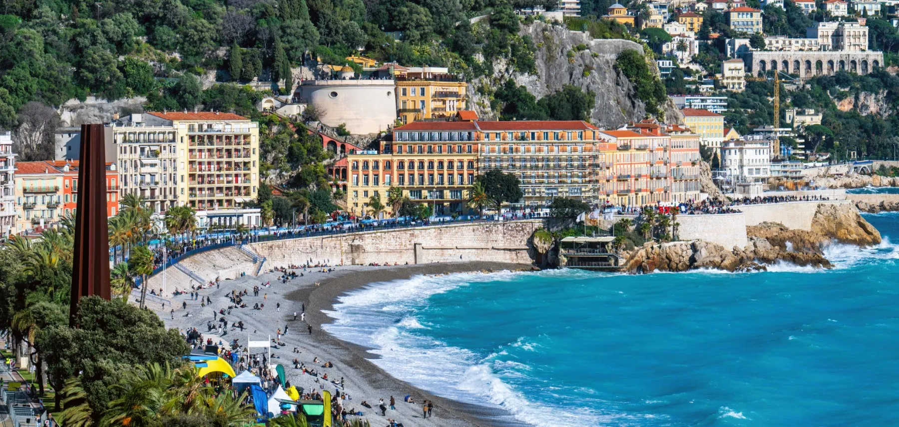 Apartment buildings along the Nice seafront below the Colline du Château, with villas climbing the wooded hillside above them and people on the pebble beach below