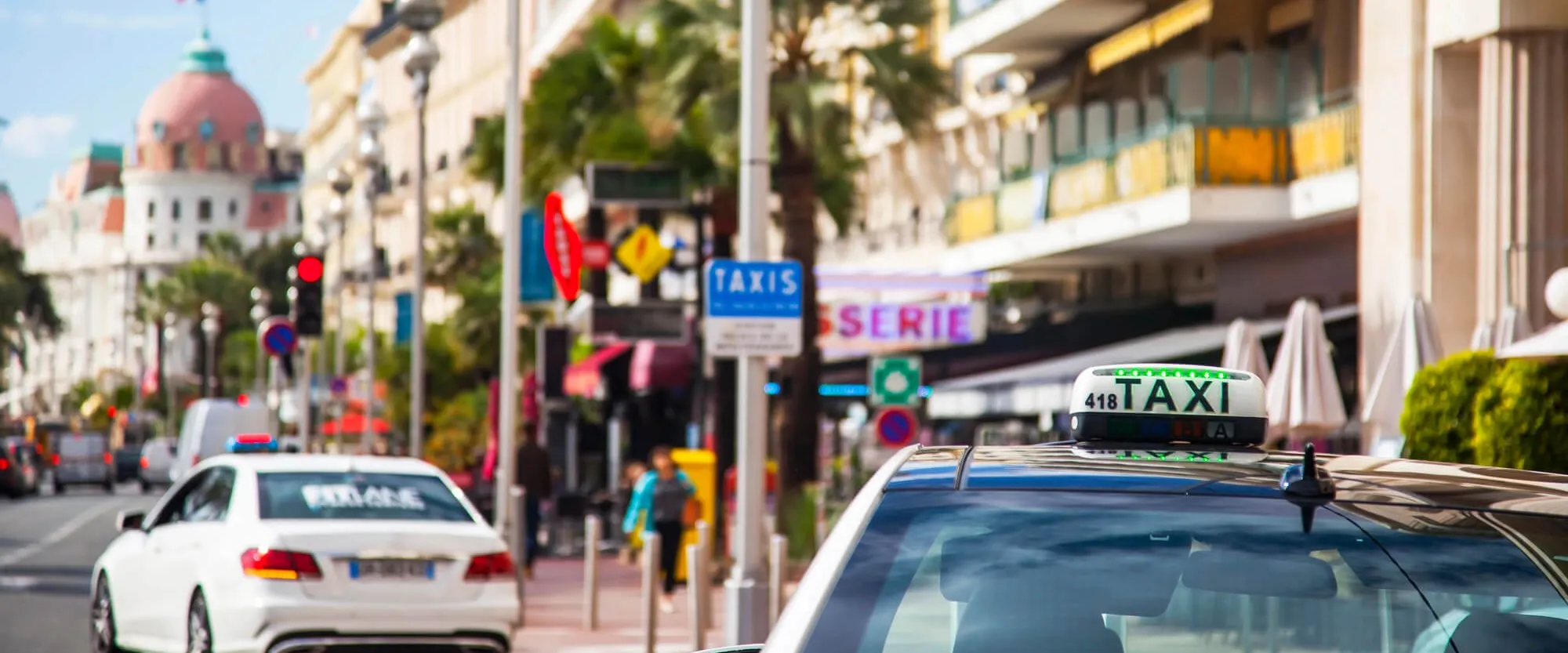 A taxi on the Promenade des Anglais in Nice with its rooftop sign lit on tariff A, a blue TAXIS rank sign and the pink dome of the Hôtel Negresco behind it