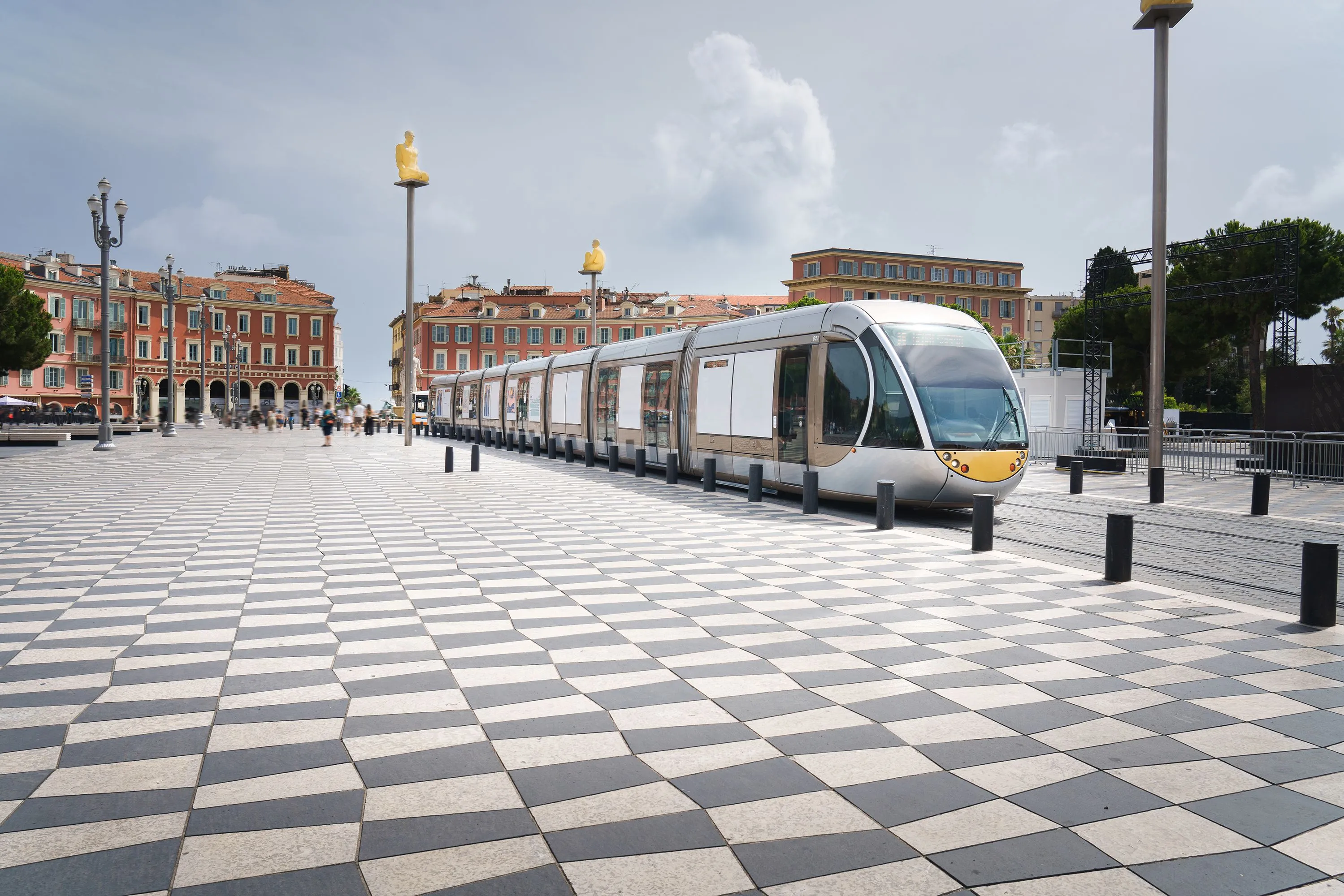 A modern tram crossing the checkered pavement of Place Masséna in Nice, with the city's yellow Conversation à Nice statues in the background