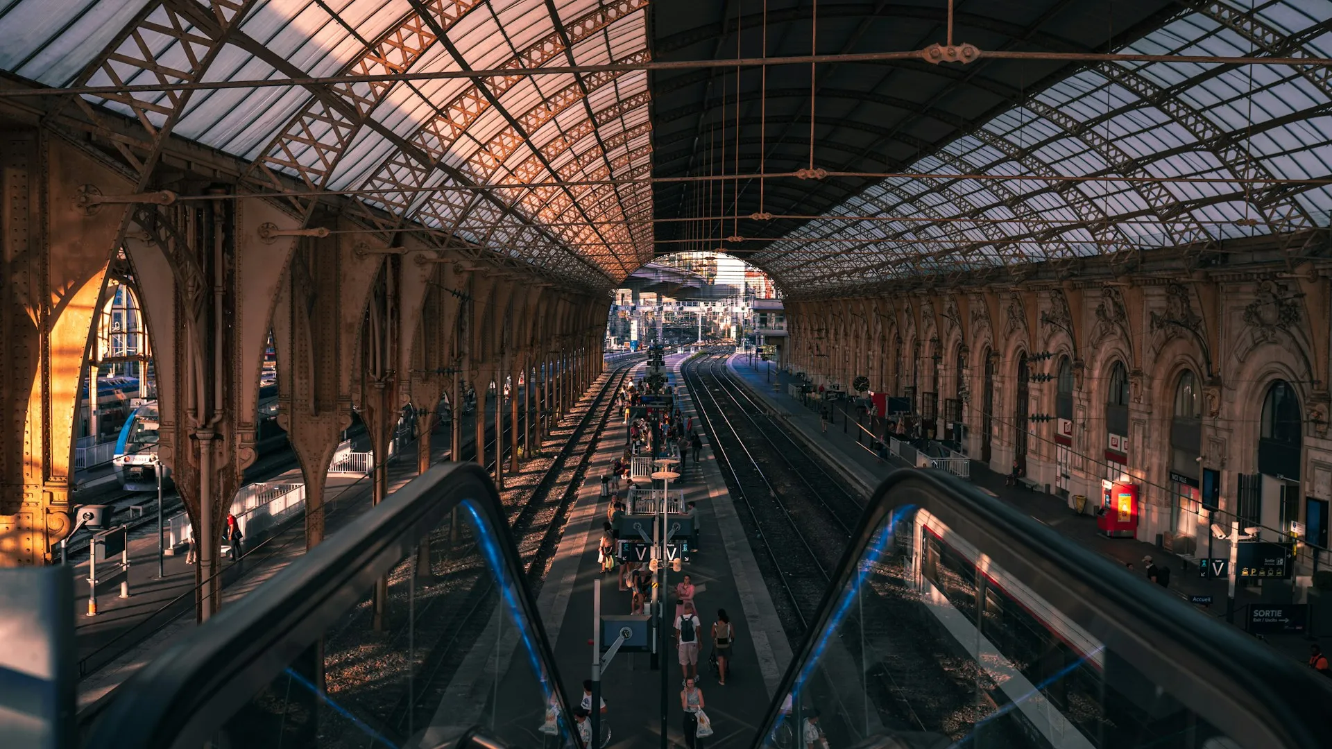 The platforms at Nice-Ville station seen from above in evening light, with the arched glass and ironwork canopy overhead, passengers walking along the platform and a regional train waiting at the far side