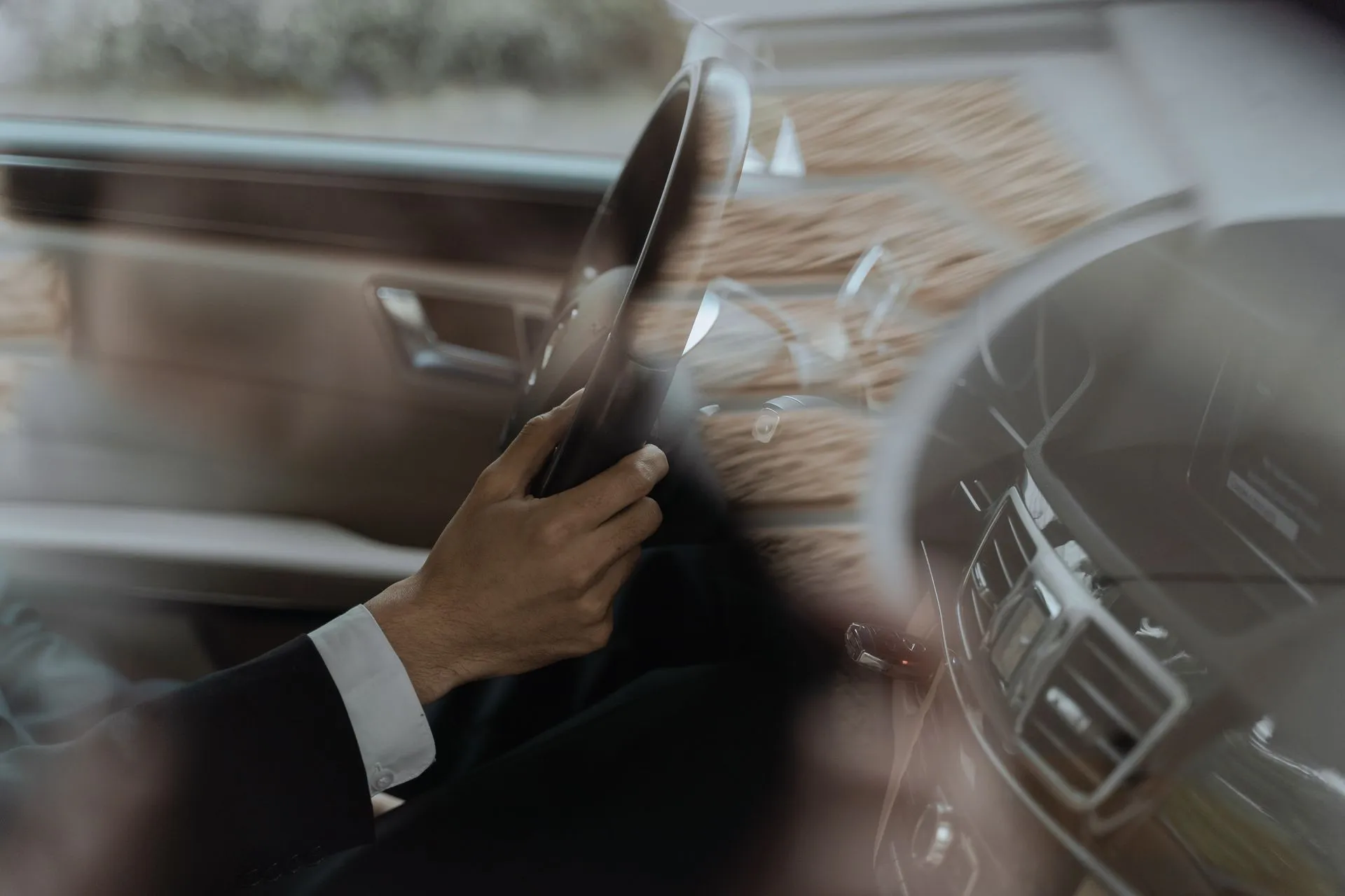 A chauffeur in a dark suit and white shirt cuff driving an executive car, one hand on the steering wheel, the landscape blurring past the window