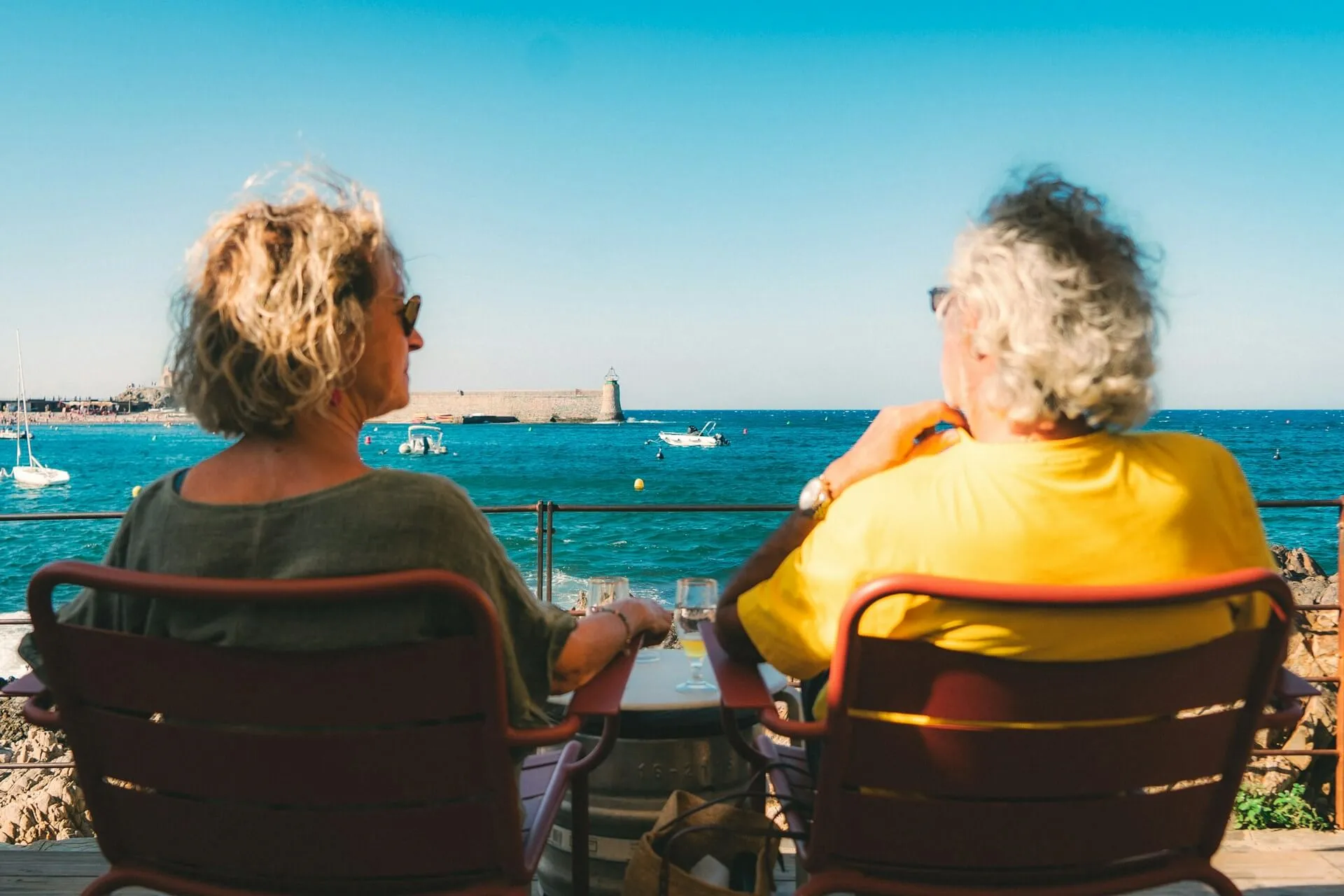Two older people sitting at a seafront table on the Côte d'Azur, looking out over turquoise water toward a stone fort and small boats at anchor