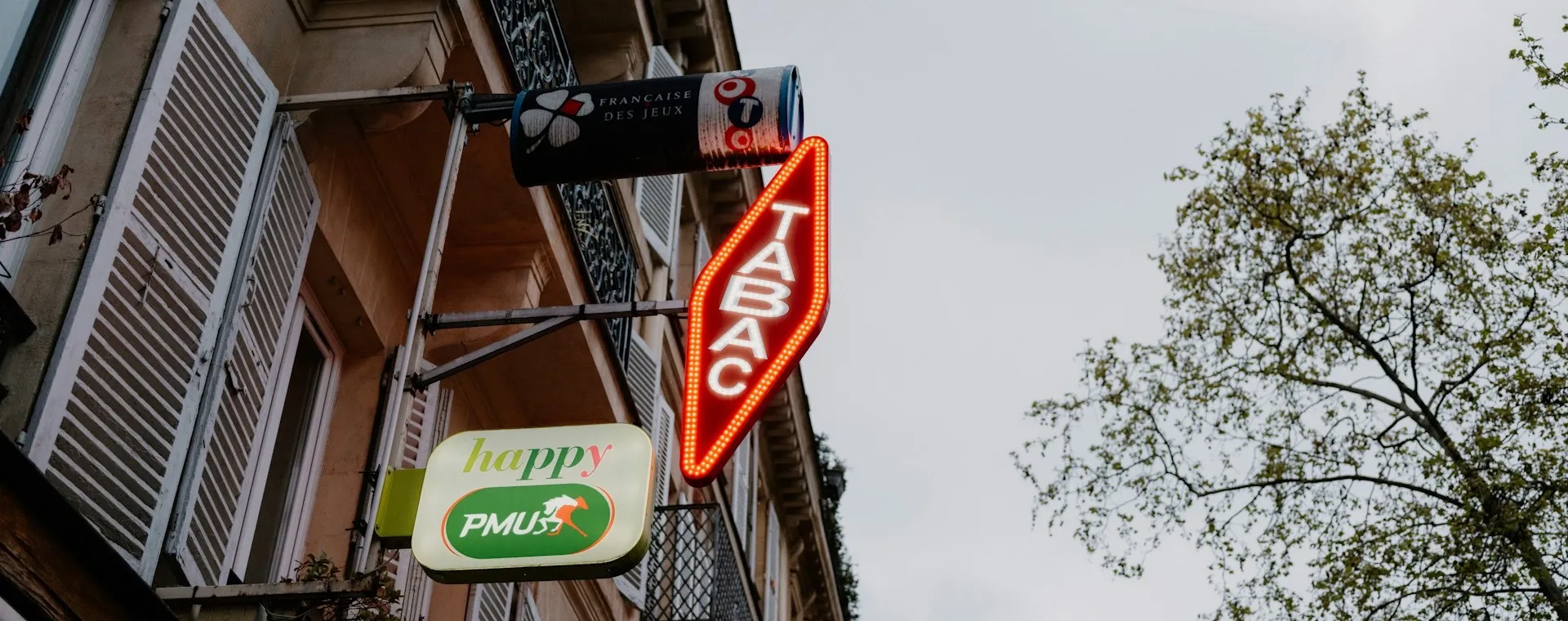 A lit red diamond-shaped TABAC sign on a French apartment building, next to a Française des Jeux sign and a green PMU sign above a newsagent