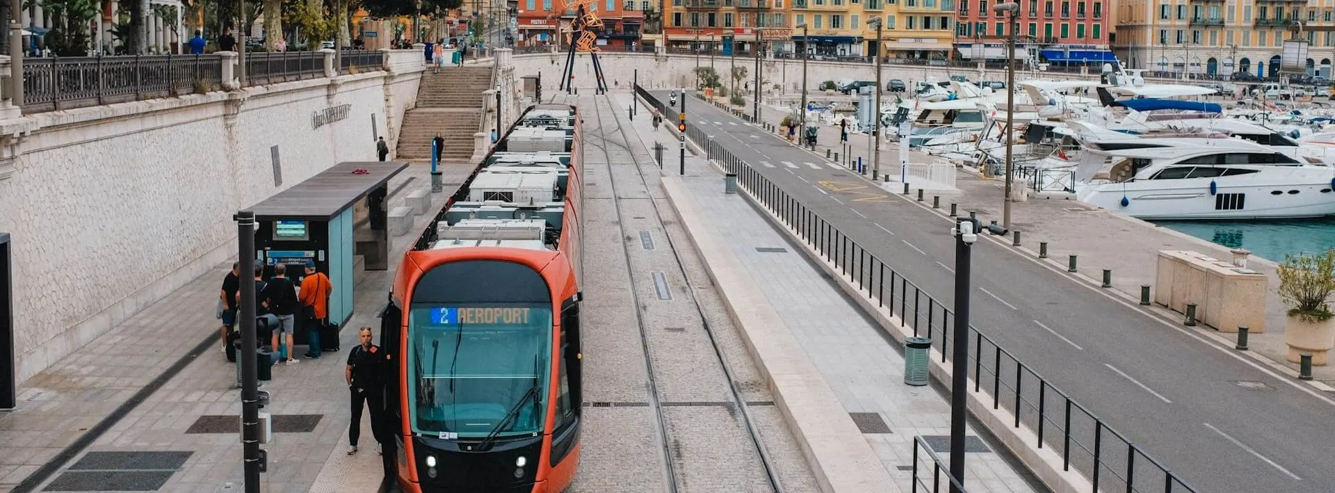 A red tram line 2 waiting at a stop beside Nice's port, its destination board reading 2 AEROPORT, with passengers and luggage on the platform and yachts moored across the quay