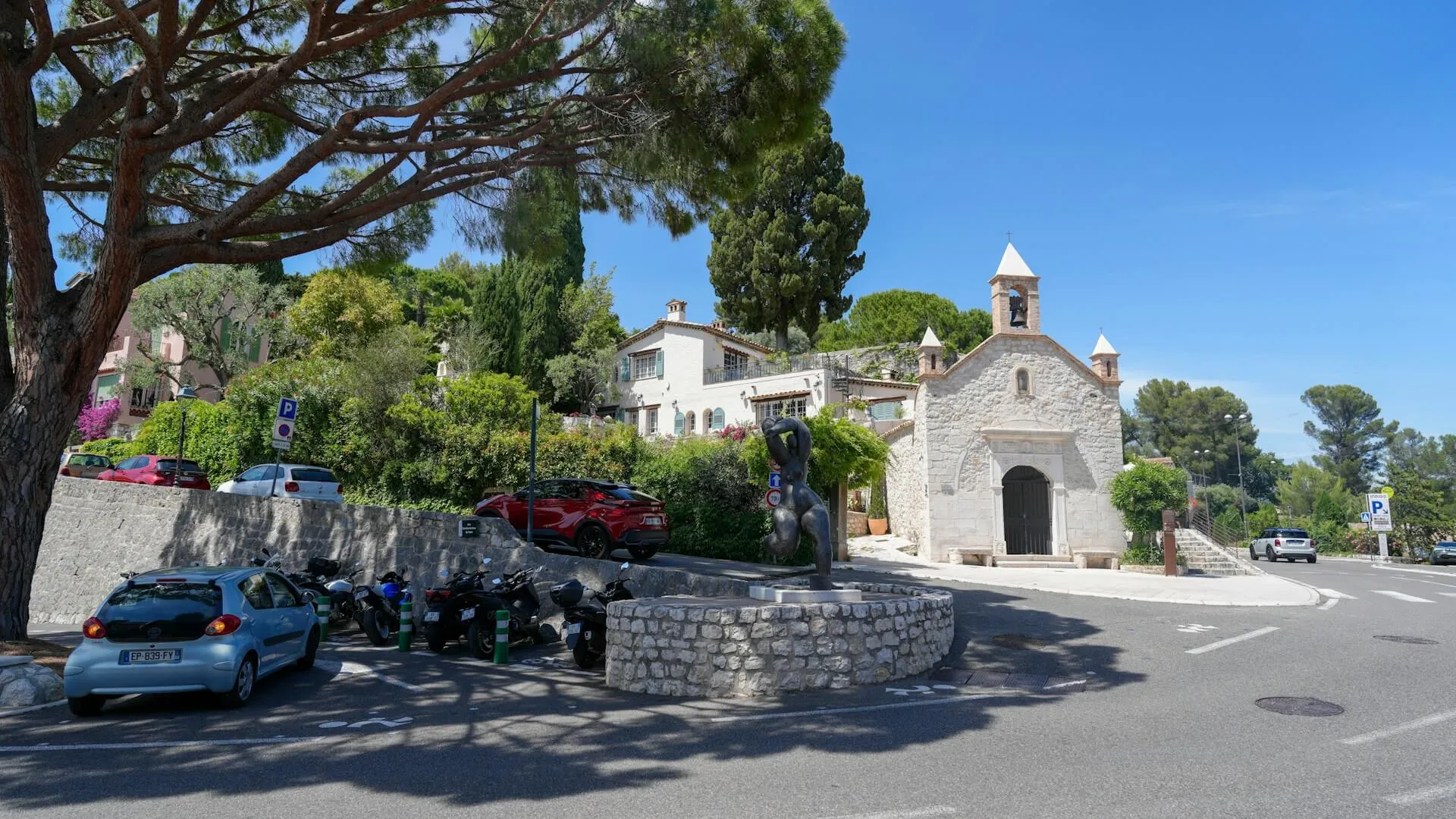 Cars and scooters parked along a Côte d'Azur village square beside a small stone chapel, with blue parking signs and a road curving away under umbrella pines